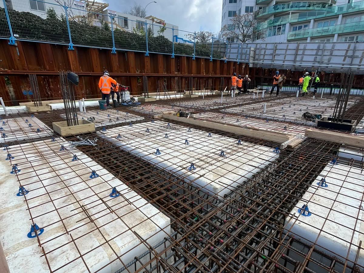 Reinforcing steel and void formers laid out across a raft slab inside a sheet piled excavation, apartment tower behind