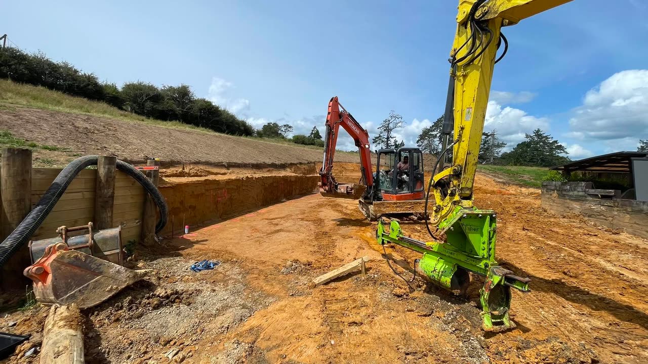 Excavator mounted post driver setting timber poles for a retaining wall, with a second excavator trimming the cut behind
