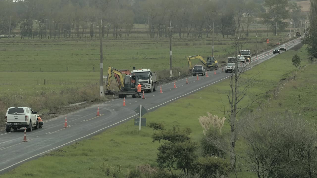 Excavator and tipper truck working the shoulder of a rural highway behind a cone taper, with traffic queued past the site