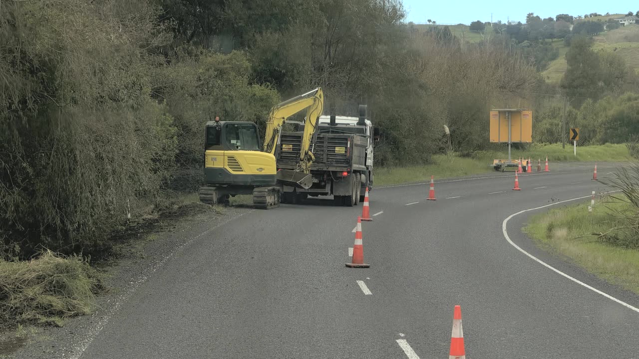 Excavator loading spoil into a tipper truck on the road shoulder with cones and a variable message board
